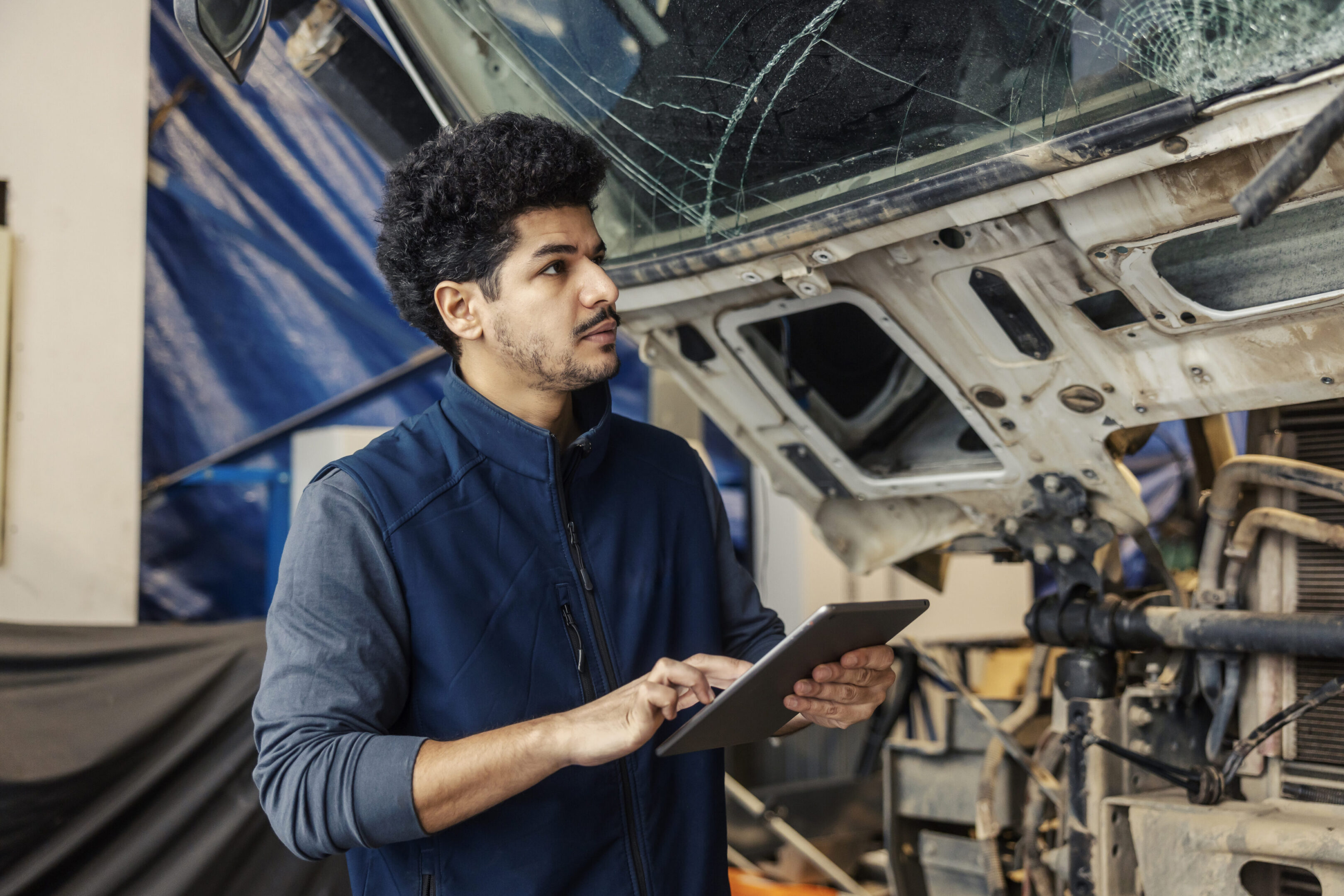 Serious diverse technician assesses the damage with tablet on truck made in an accident at mechanic workshop.